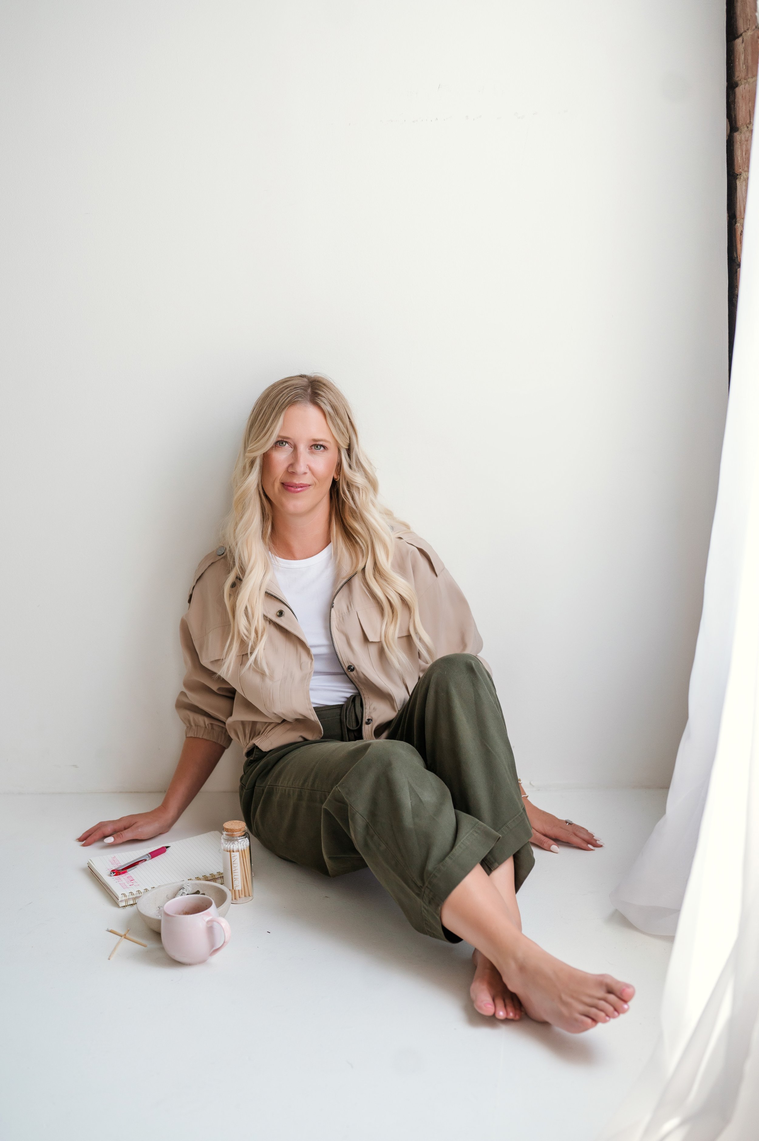 A woman with long blonde hair, wearing a beige jacket and green pants, sitting on the floor against a white background, looking at the camera with a slight smile. There are a notebook, pen, a jar of vitamins, a pink mug, and a spoon on the floor beside her.