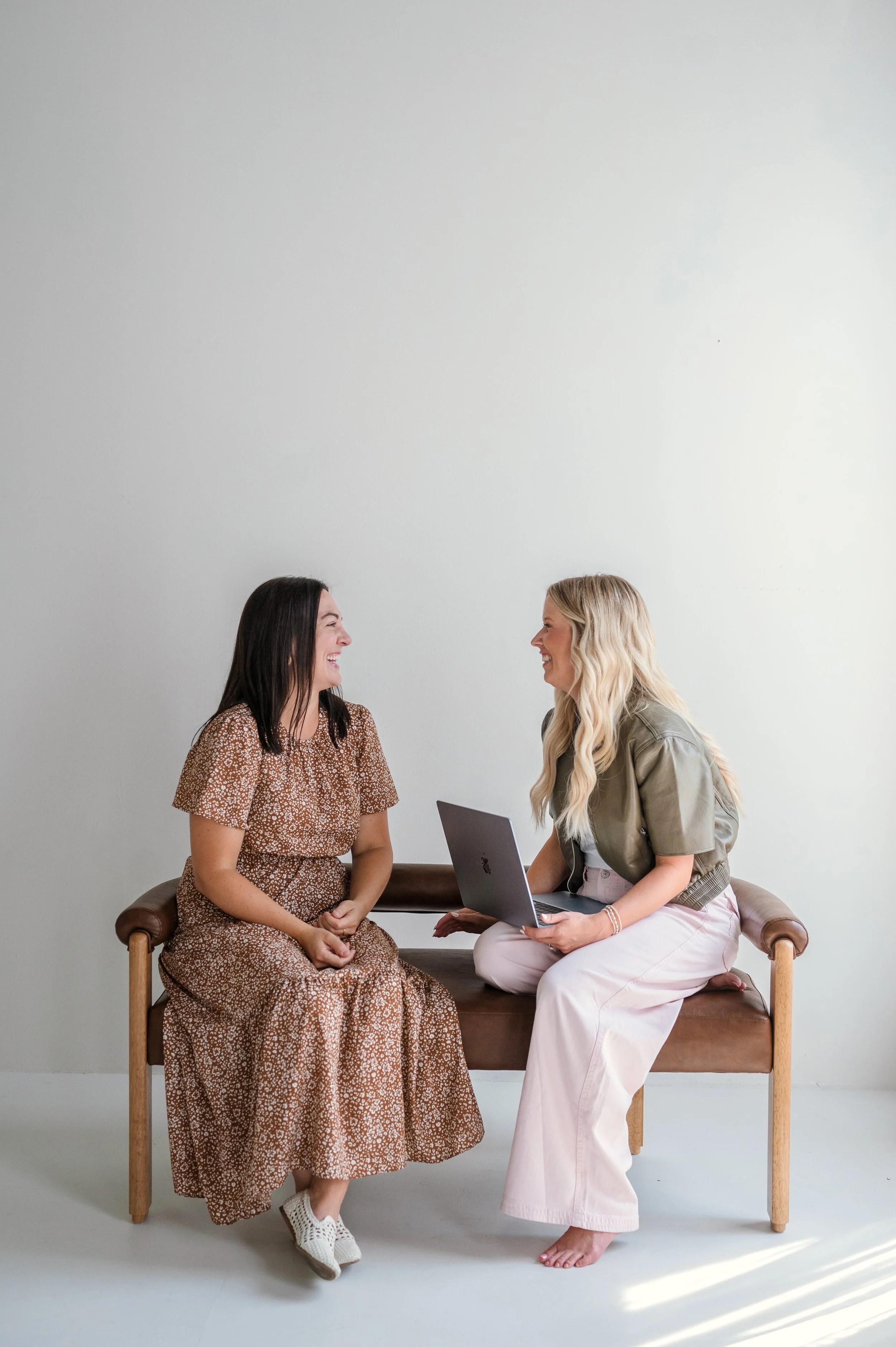 Two women sitting and chatting on a wooden bench against a plain white wall; one is holding a laptop, and they are smiling at each other.