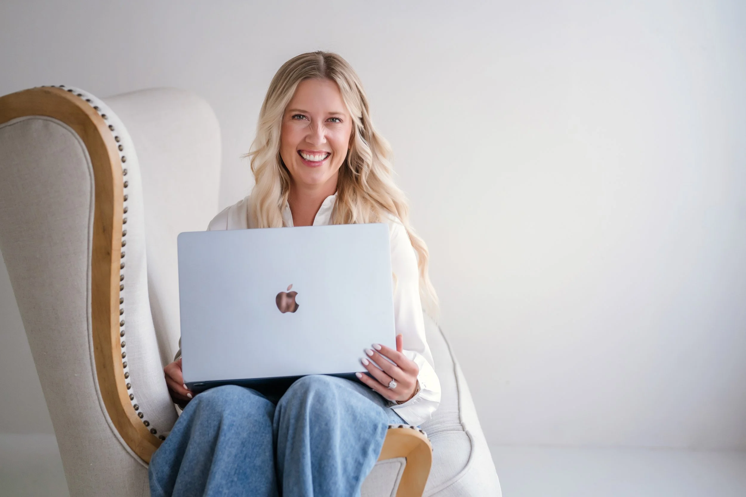 A woman sitting on a beige armchair with a padded back, holding a silver MacBook laptop, smiling at the camera, against a plain white wall.
