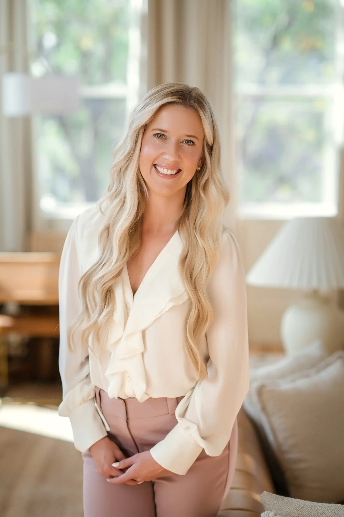 A woman with long blonde hair, smiling, wearing a cream blouse with ruffles and pink pants, standing in a sunlit living room with large windows and beige decor.