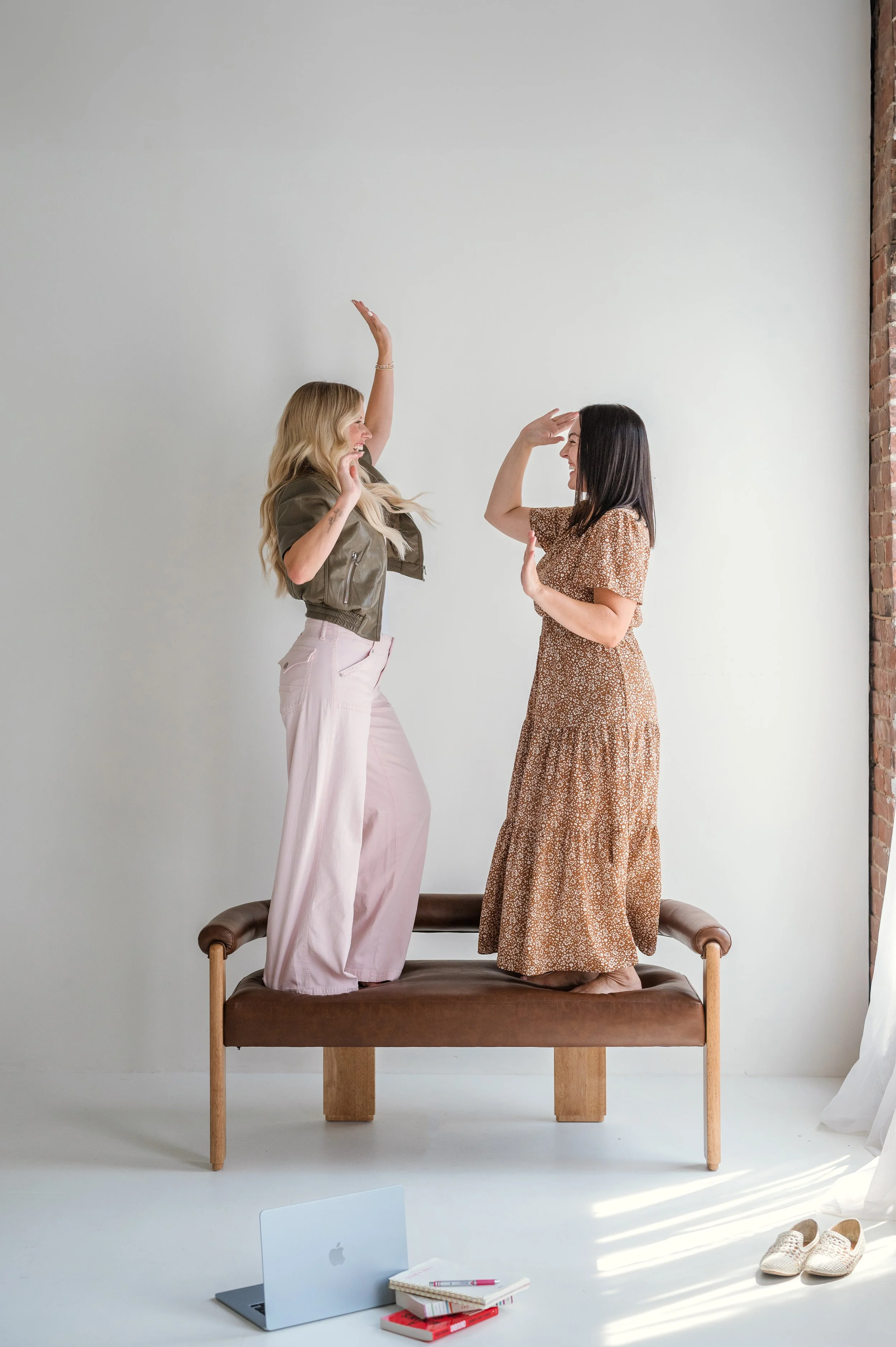 Two women are standing on a padded bench, facing each other and smiling. They are reaching out to high-five each other with one hand while the other hand is on their foreheads in a salute gesture. One woman has long blonde hair, wears a brown jacket and light pink pants, and the other woman has black hair, wears a brown patterned dress. The background is a plain light grey wall with an exposed brick section on the right. There are shoes on the floor and a laptop, notebooks, and pens on the floor below the bench.