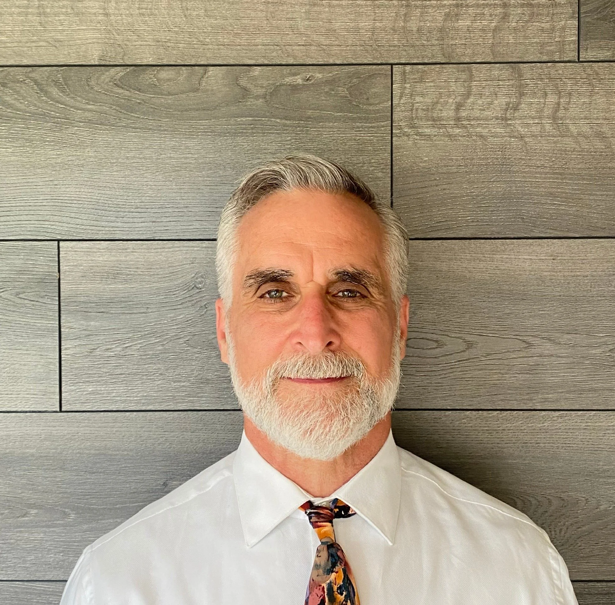 Close-up of a smiling middle-aged man with gray hair and beard, wearing a white shirt and a colorful tie, standing in front of a wooden wall.