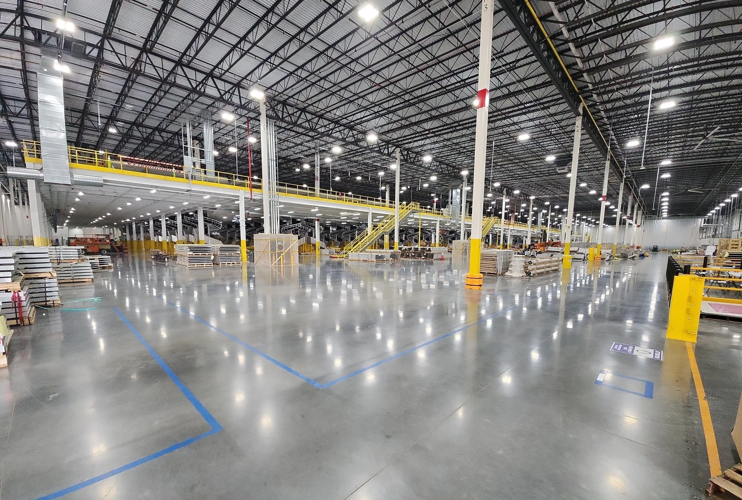 Empty warehouse with high ceiling, metal beams, bright overhead lights, yellow safety railings, stairs, and various equipment and pallets on the floor.