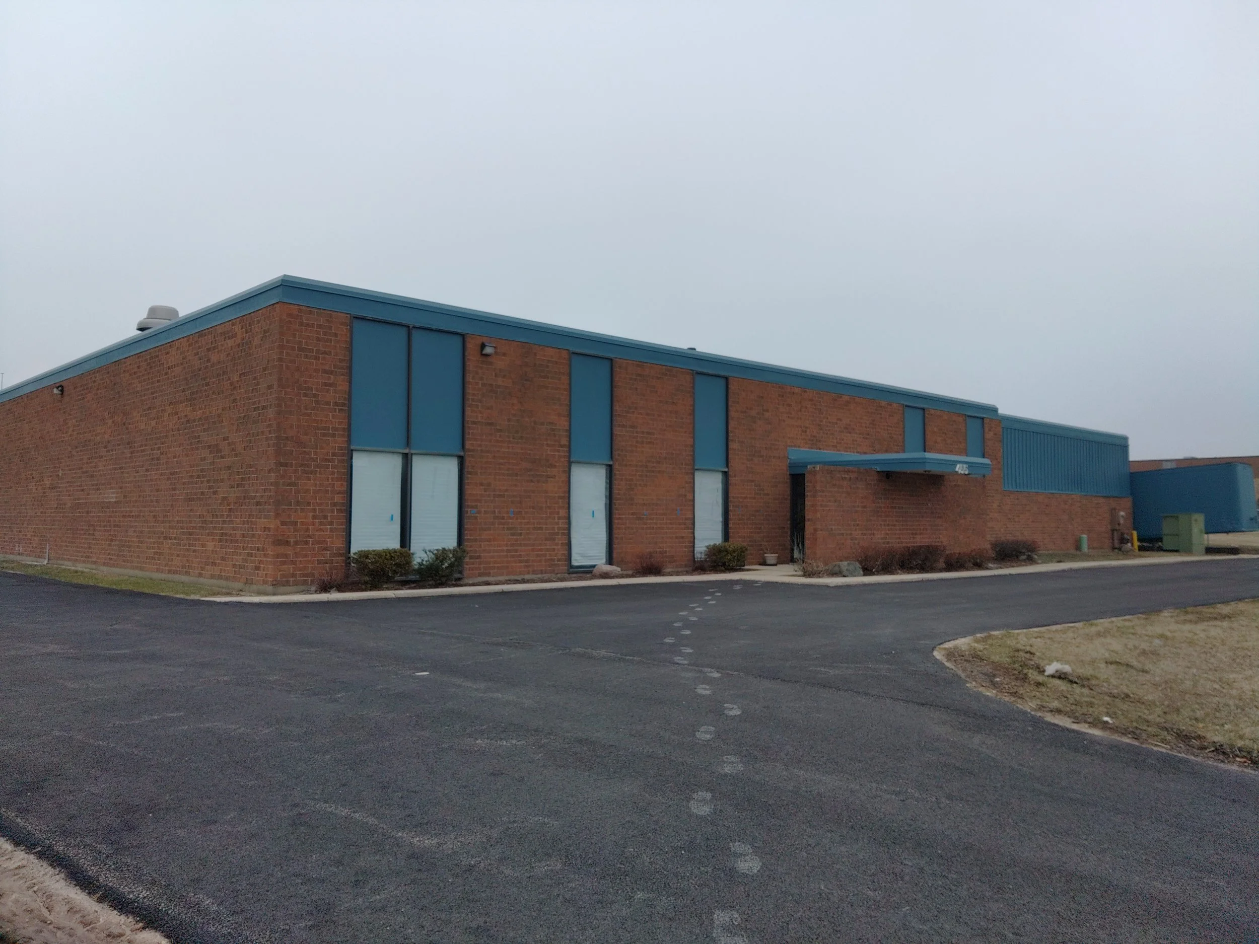 Photo of a brick building with blue accents and windows. The building is next to a parking lot with a curved driveway and some small bushes.