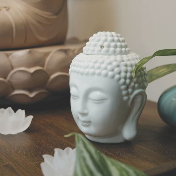 White ceramic Buddha head surrounded by soft neutral decor, greenery, and white flowers.