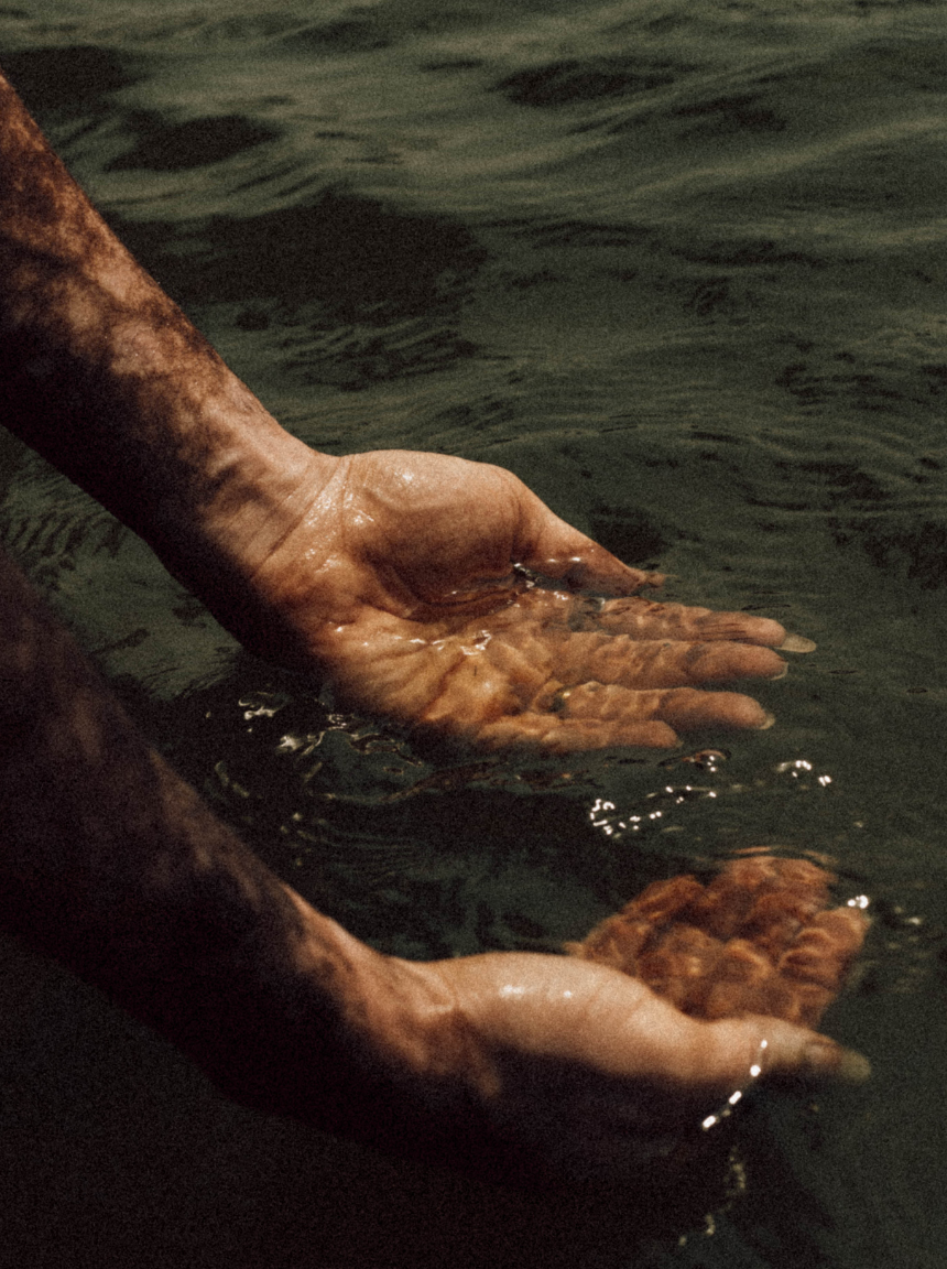 A person with tanned, wet arms is holding a large, reddish-brown rock underwater in a body of water.