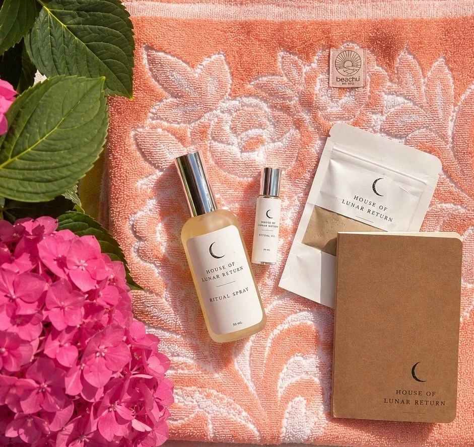 Ritual products from House of Lunar Return, including a ritual spray, oil, and a packet, arranged on a floral textured cloth with pink hydrangea flowers and green leaves nearby.