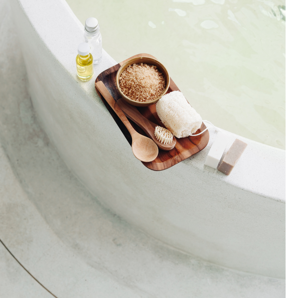 A wooden tray on the edge of a bathtub with bath salts, a loofah, a wooden body brush, soap, and bottles of oil and soap.