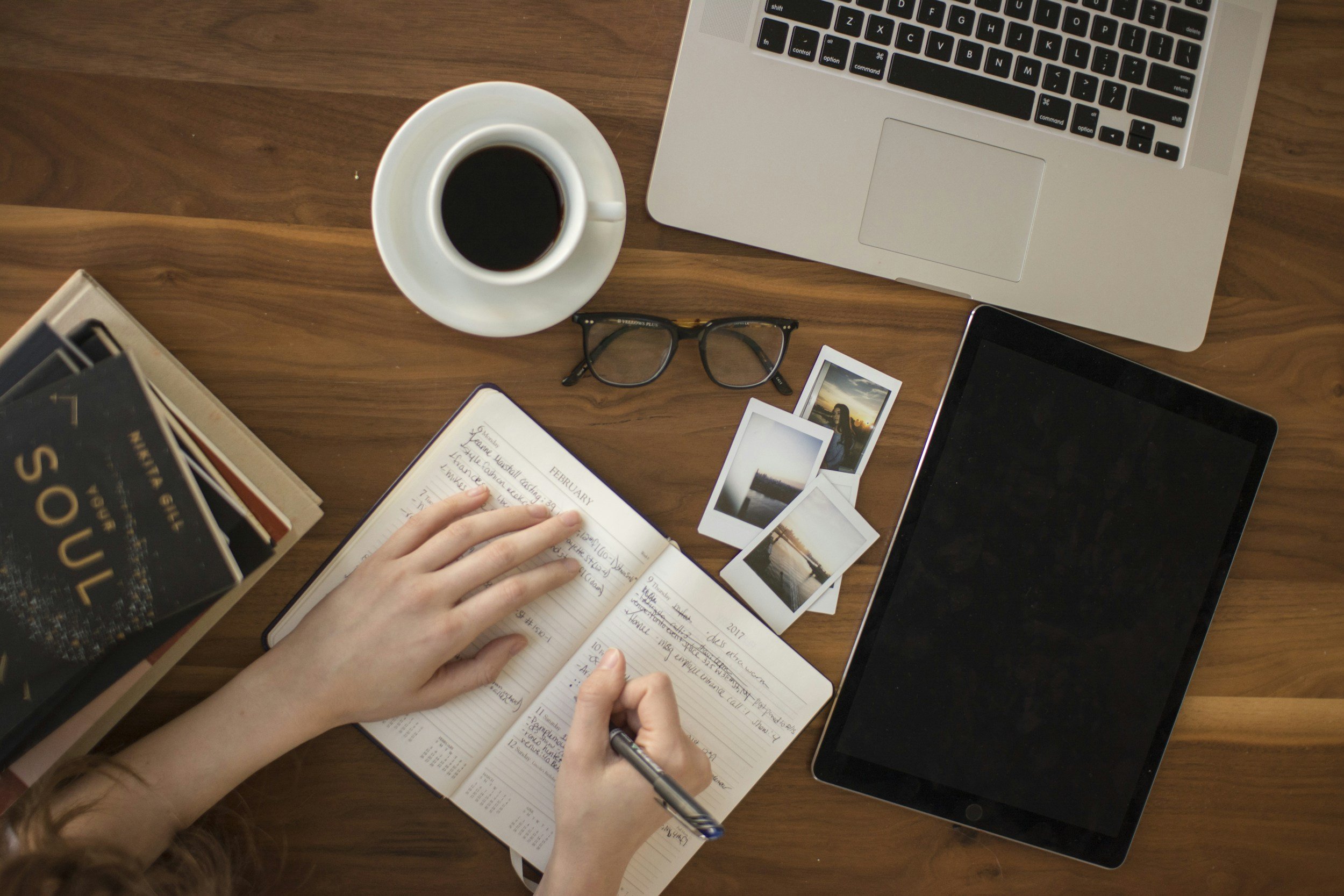 A person writing in a planner on a wooden desk, with a laptop, a cup of coffee, glasses, printed photos, and a tablet nearby.