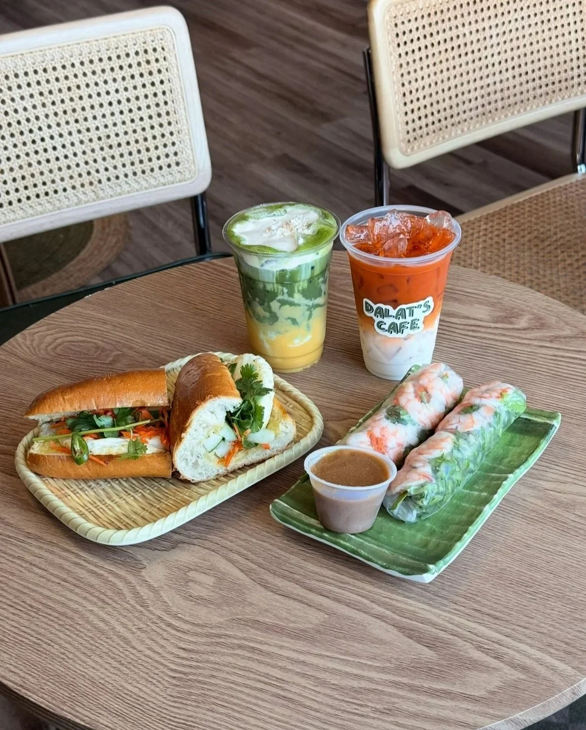 A table with Vietnamese food including a banh mi sandwich, two spring rolls with dipping sauce, and two drinks, one iced coffee and one iced tea at Dalat's Cafe.