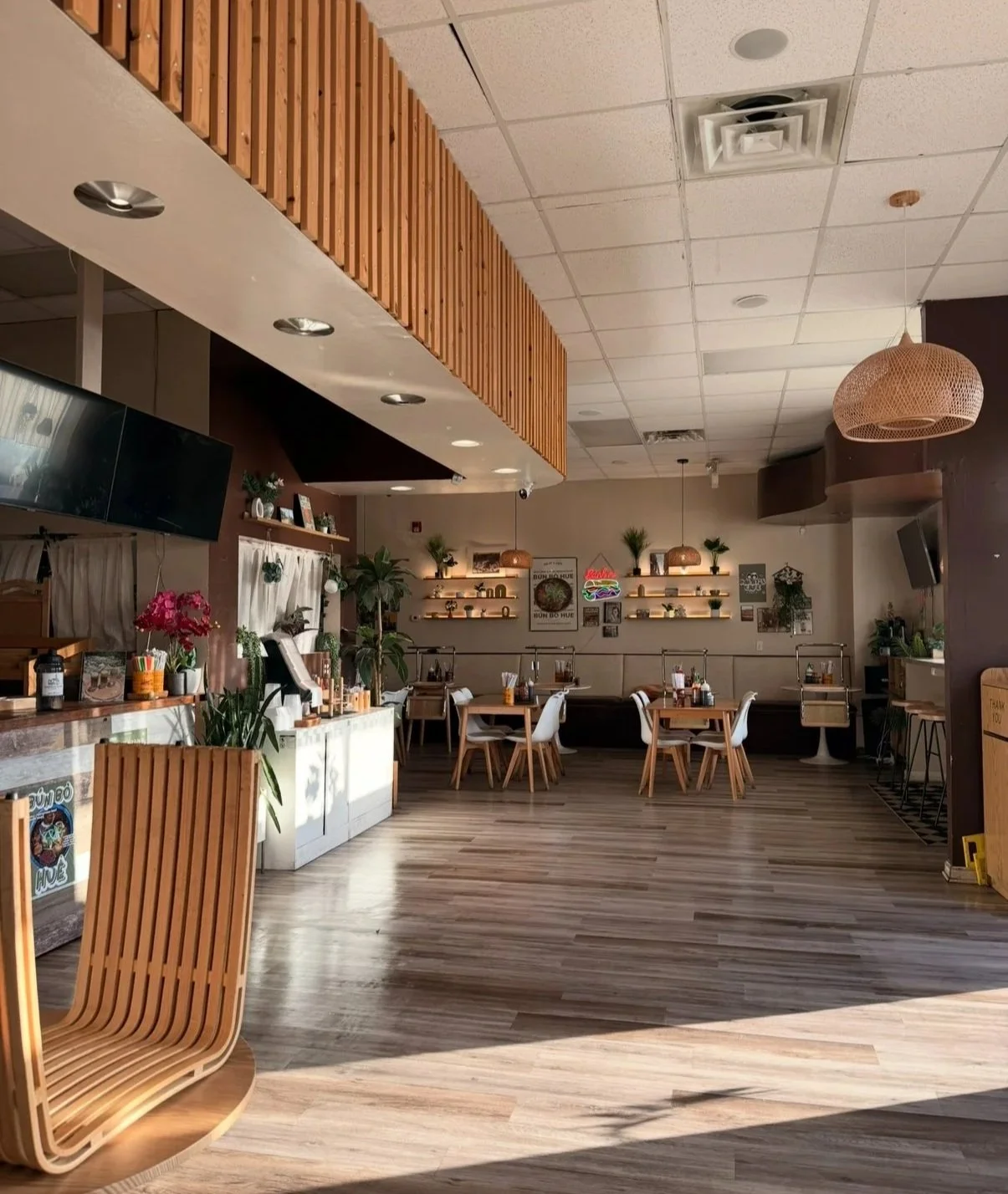 Interior of a modern, cozy cafe with wooden furniture, green plants, and decorative lighting fixtures. There are tables and chairs arranged for dining, with some plants and framed pictures on the walls.