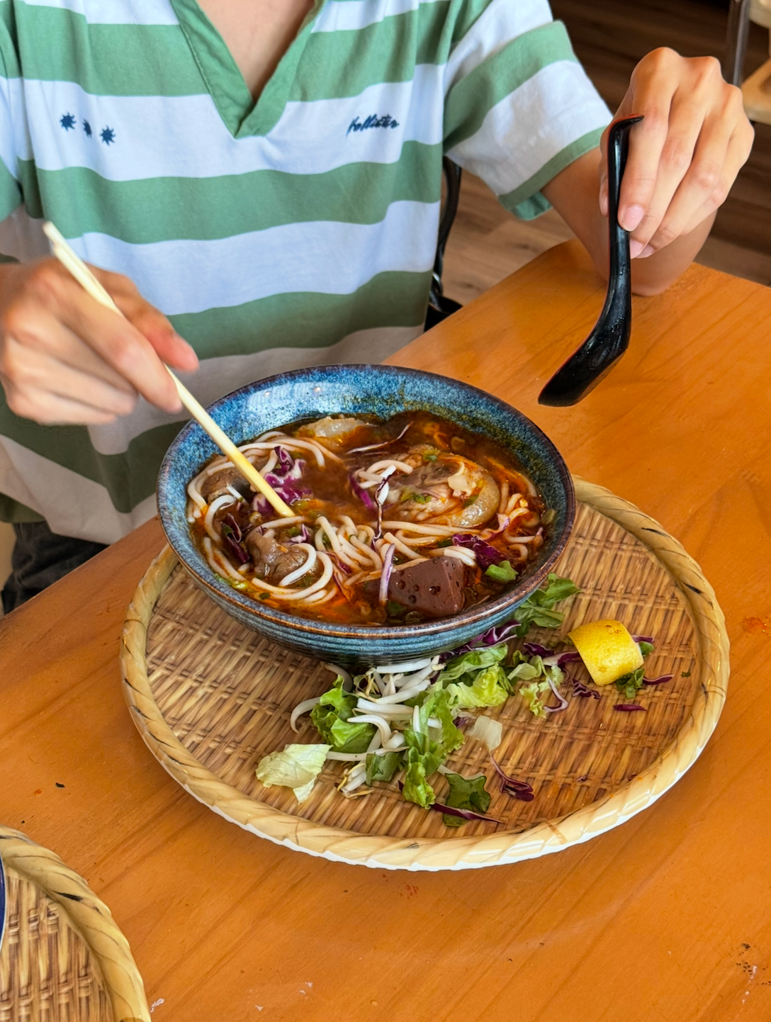 Person eating a bowl of Asian noodle soup with chopsticks at a wooden table, with a decorative woven tray with a lemon and salad greens on the side.