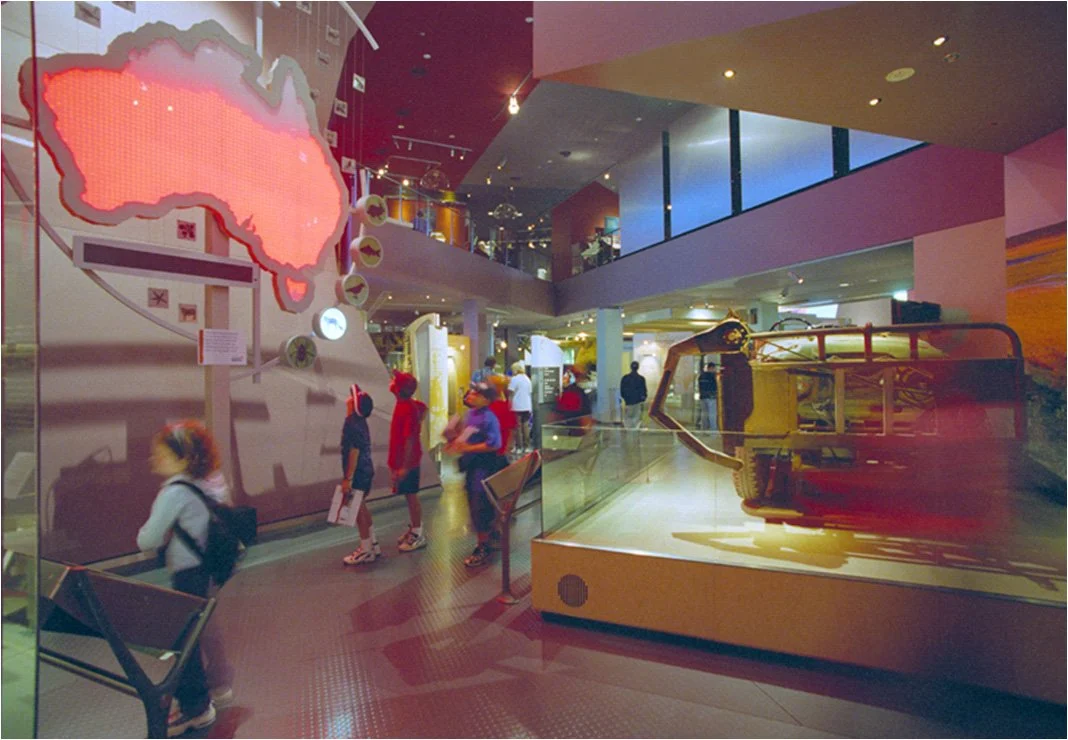 Interior of a museum with children exploring exhibits, including a large map of Australia and a display case with a mechanical excavator.