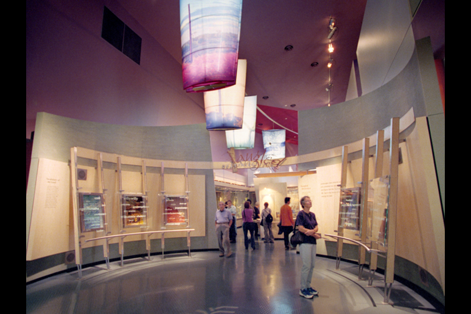 At the National Museum of Australia, a Museum exhibit featuring informational panels on a curved display wall, with visitors observing and reading the displays, and colorful hanging fabric art installations overhead.