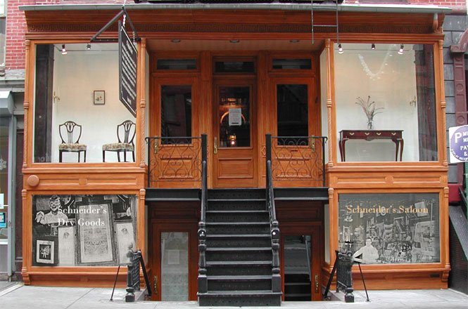Front view of a two-story wooden storefront with stairs leading to an entrance door, large windows displaying chairs and a console table, and black and white photographs on the lower panels.