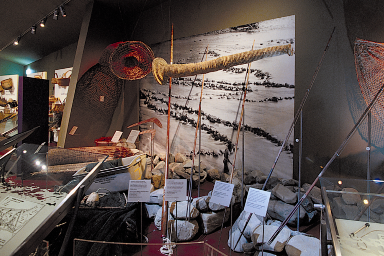 At the National Museum of Australia, a Exhibit with fishing equipment including nets, poles, and a boat, displayed on a rocky area against a backdrop of a large black and white landscape painting.