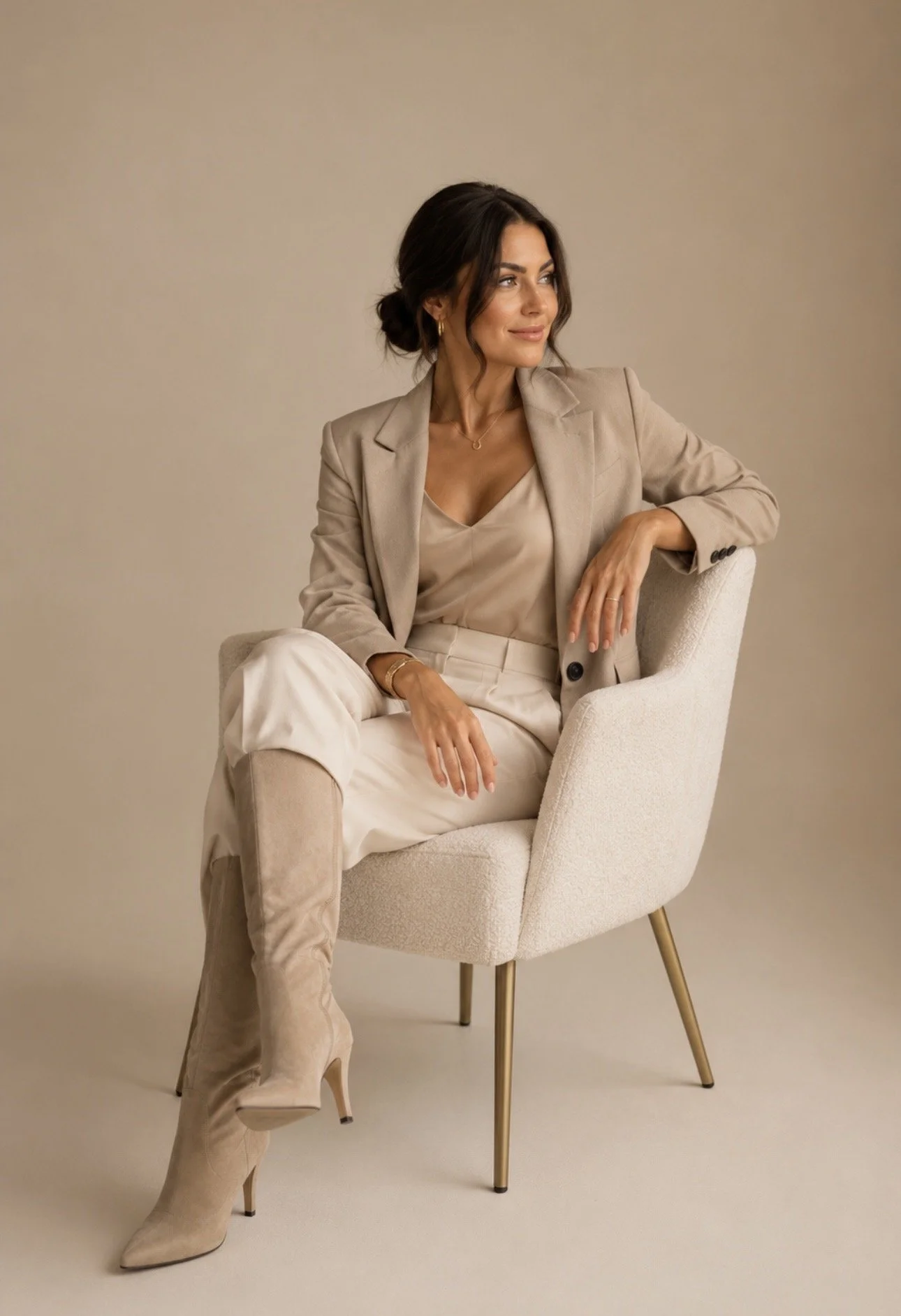 Woman in beige blazer and pants, sitting in a cream-colored armchair against a plain beige background.