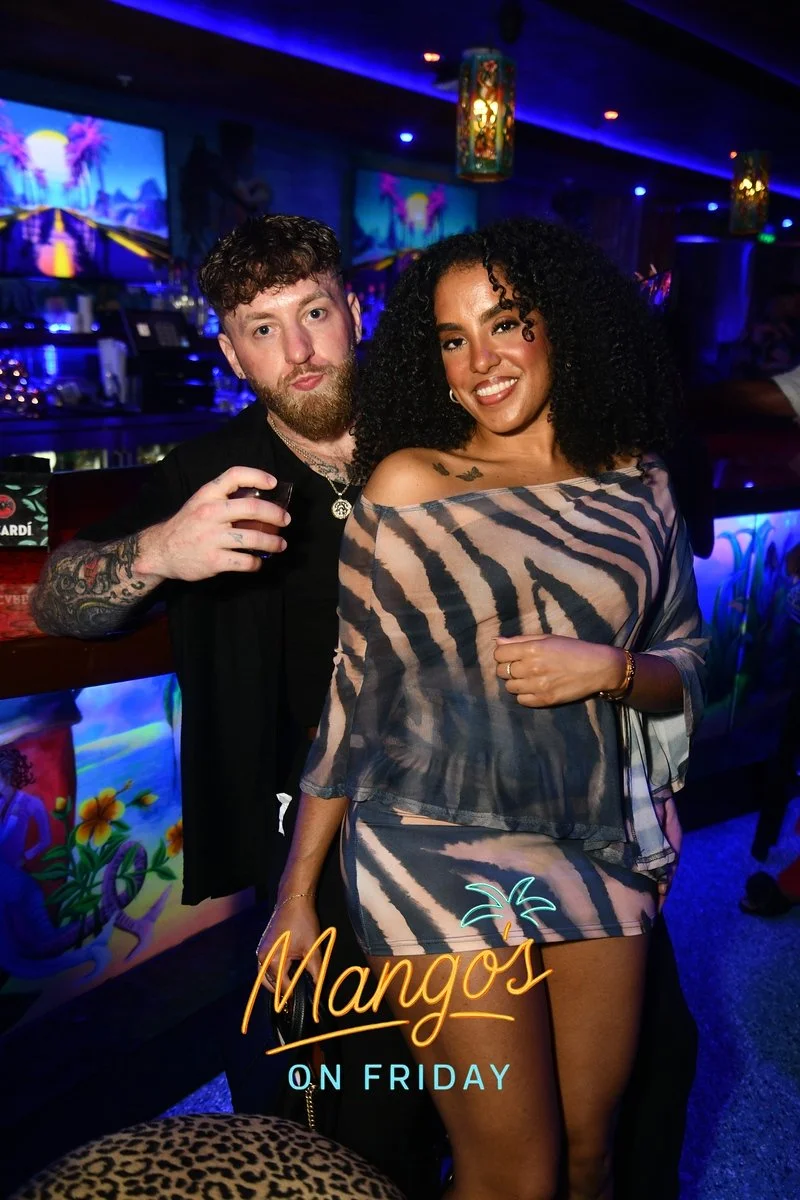 A man and woman smiling at a bar in a nightclub with neon lights, tropical-themed decor, and a responsive display that reads 'Mangos on Friday'.