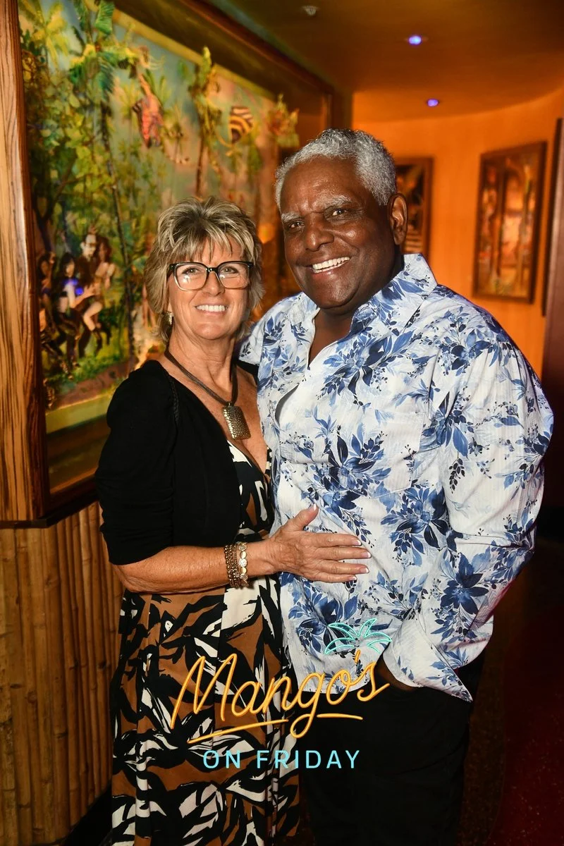 A smiling woman with glasses and blonde hair, and a smiling man with gray hair, stand close together inside a tropical-themed restaurant or bar with warm lighting. The woman is wearing a patterned dress and a black cardigan, while the man is wearing 