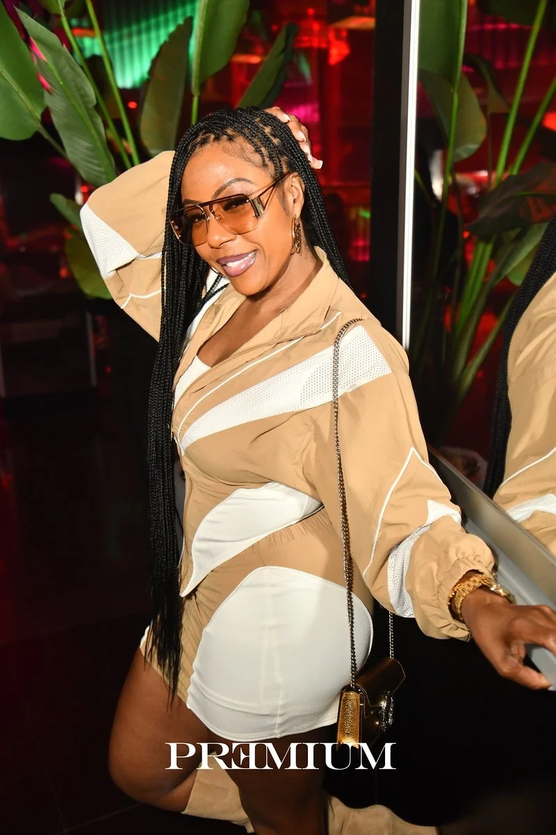 A woman with long braided hair wearing beige and white sporty outfit, sunglasses, and gold jewelry, posing with one hand on her head and smiling in a vibrant indoor setting with green plants and colorful lighting.