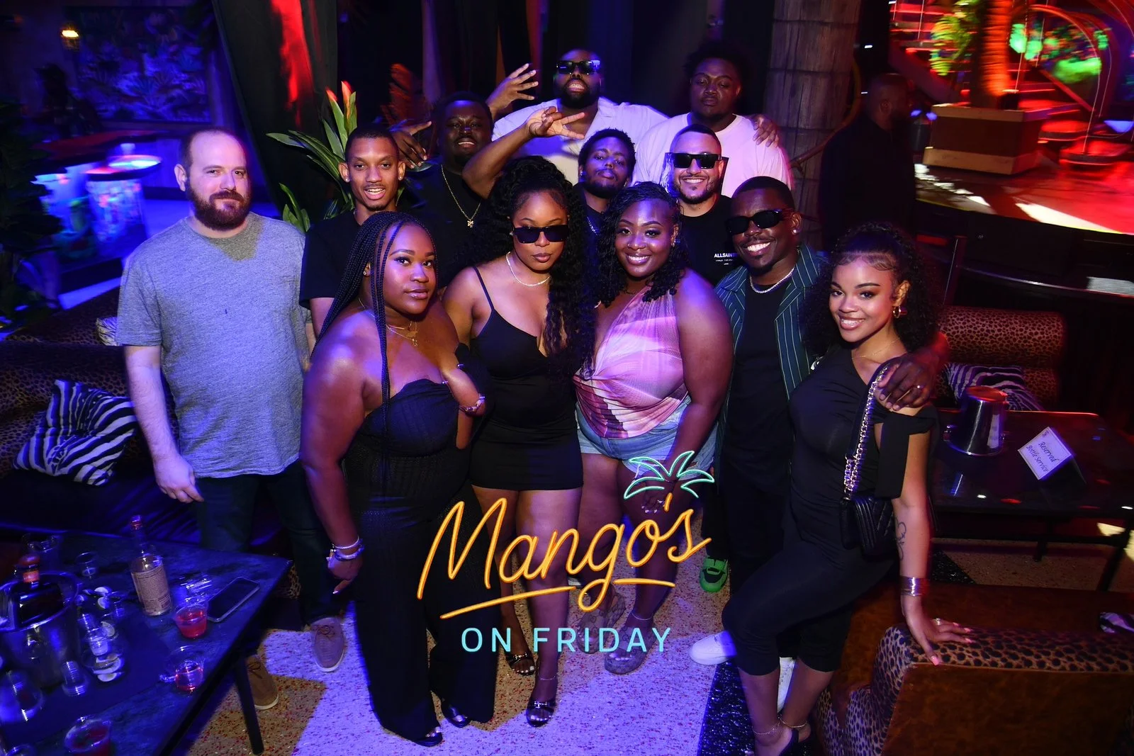 A diverse group of men and women gathered together in a lively, colorful nightclub or lounge, posing for a group photo with drinks on a table nearby, under vibrant red, blue, and green lights. The scene captures a celebratory atmosphere.