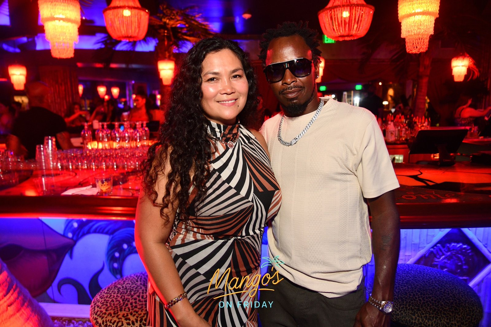 A woman and a man posing together at a bar or nightclub with red and purple lighting. The woman has long curly black hair and is wearing a sleeveless, black and white striped dress. The man is wearing sunglasses, a beige t-shirt, and jewelry. There a