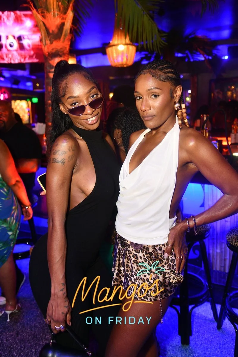 Two women with dark skin posing at a nightclub with blue and purple lighting, palm tree decor, and a neon sign in the background. One woman is wearing a black sleeveless dress and sunglasses, while the other is dressed in a white top with leopard pri