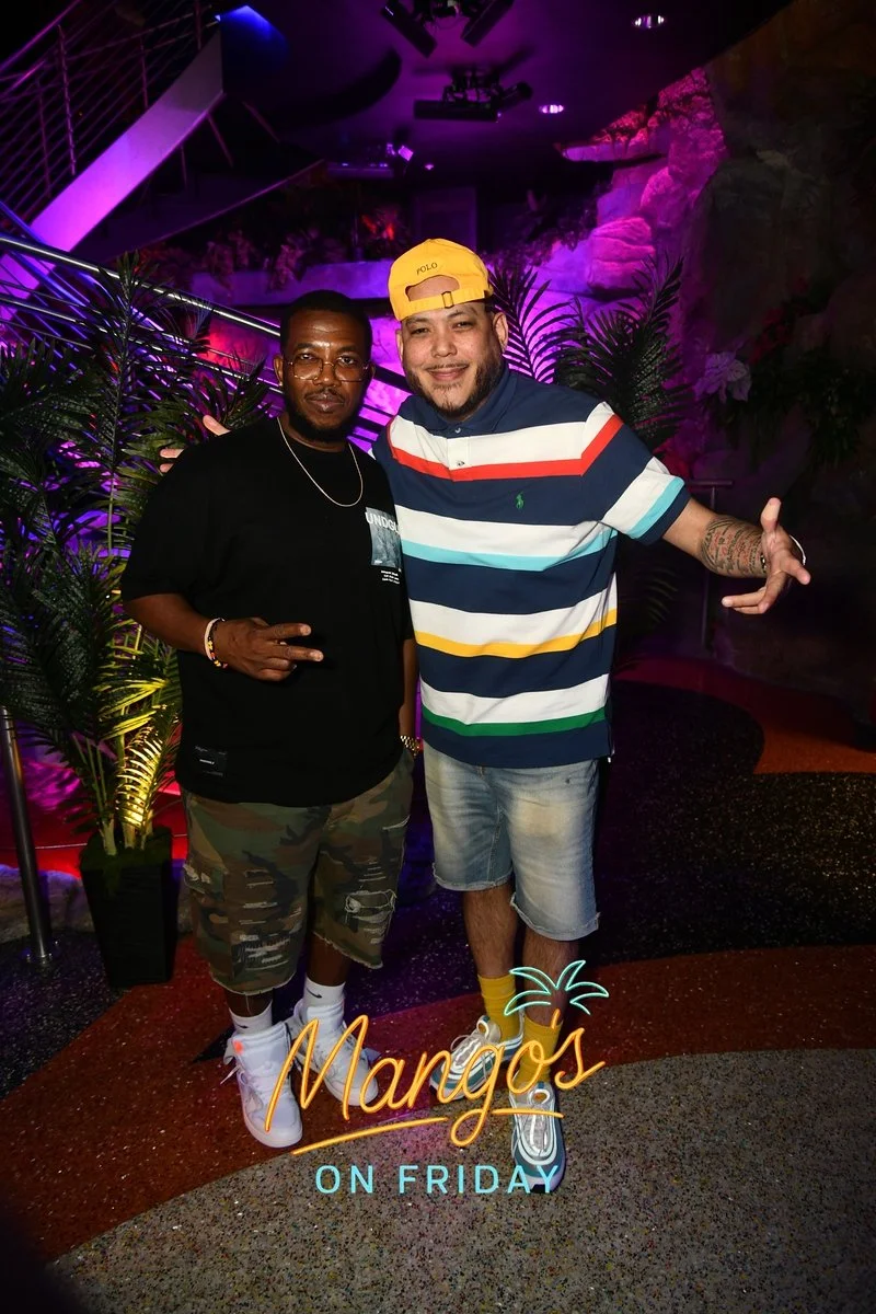 Two men standing together at a tropical-themed bar or club, with palm plants and colorful lighting in the background. One man is wearing a black t-shirt and camouflage shorts, the other is wearing a multicolored striped polo shirt, denim shorts, and 