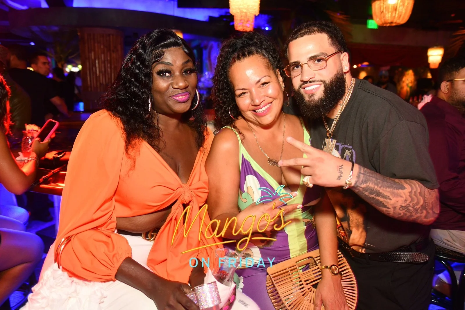 Three people smiling at a Maui's On Friday event in a lounge with vibrant lighting. One woman in an orange blouse, another in a colorful dress, and a man with tattoos, glasses, and jewelry making a peace sign.