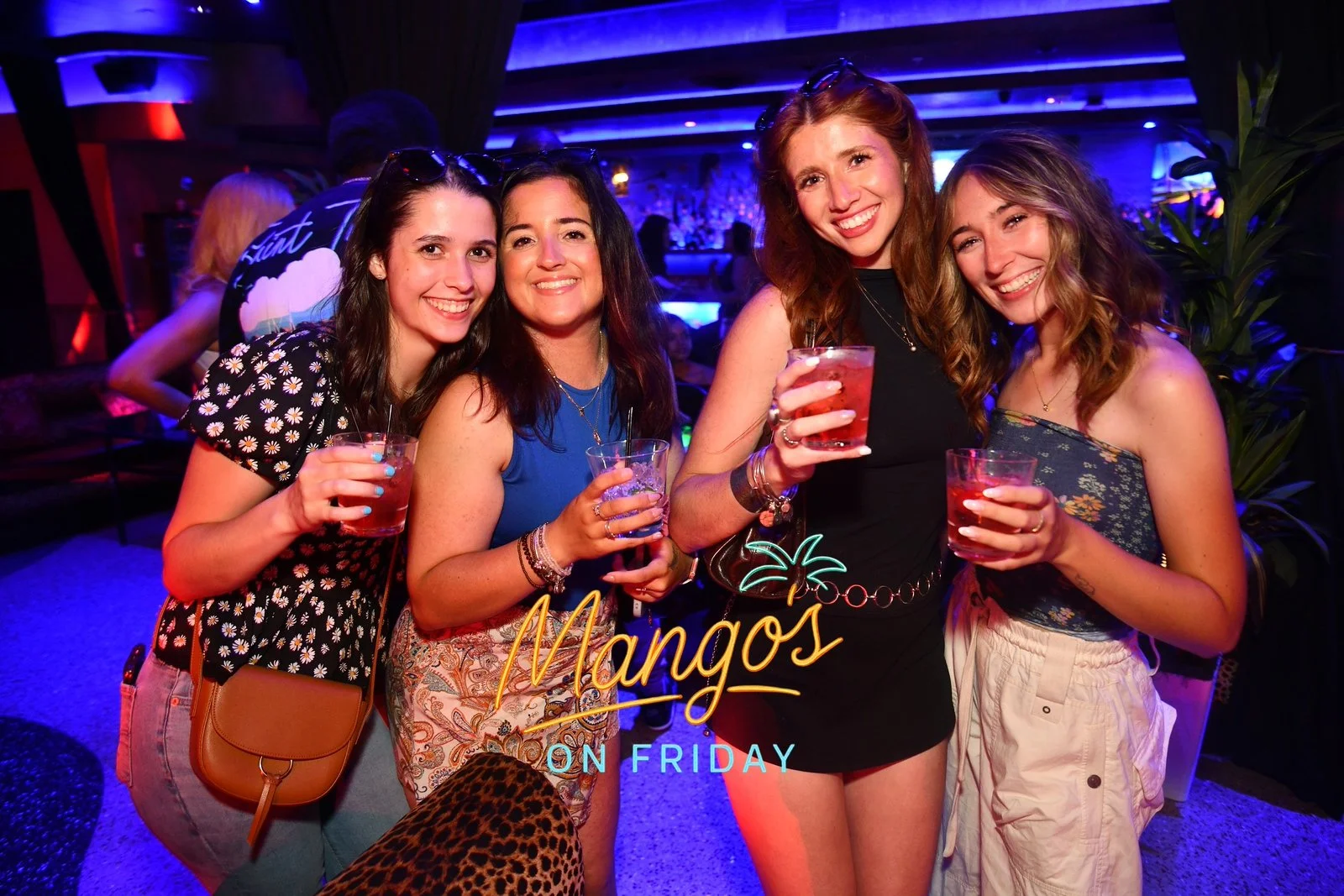 Four women smiling and holding drinks at a bar or nightclub with vibrant blue and purple lighting, and a neon sign that reads 'Mangos on Friday.'