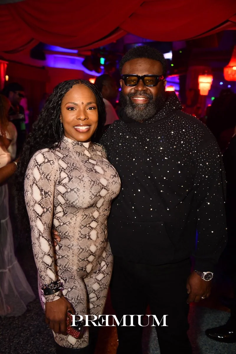 A couple posing together at a nightclub with colorful lighting. The woman is wearing a snakeskin-patterned dress and holding a phone, while the man is wearing a black hoodie with sparkly embellishments and sunglasses.