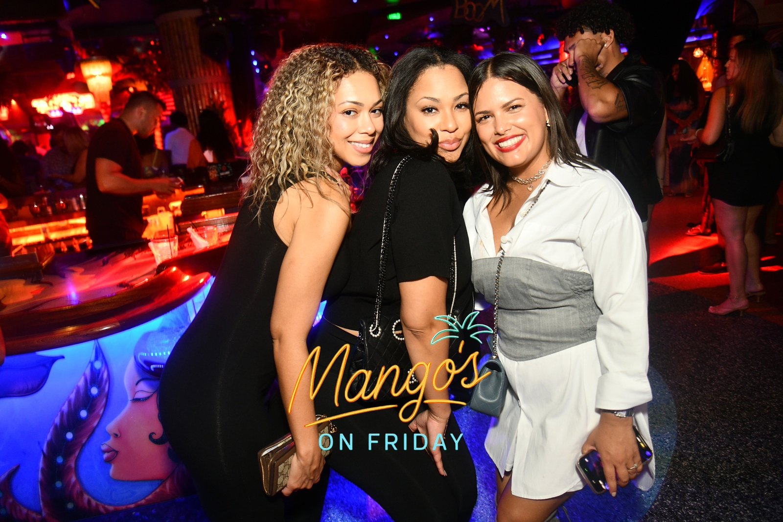 Three women smiling and posing together at a nightclub bar, with a neon sign that says 'Mango's on Friday' in front of them, surrounded by colorful lights and people in the background.