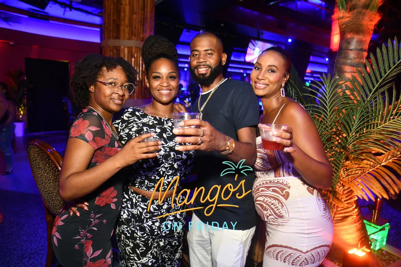 Group of four friends at a nightlife event called Mango's on Friday, holding drinks and smiling, with tropical decor and colorful lighting in the background.