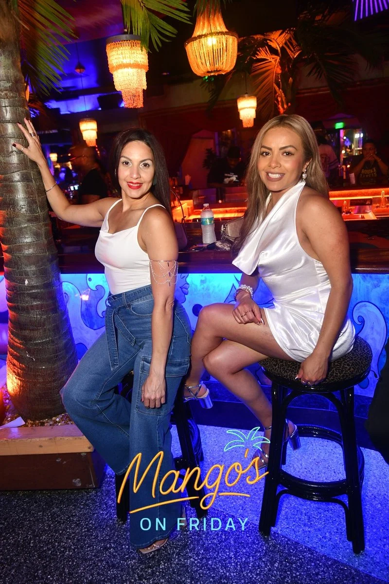 Two women posing in a dimly lit nightclub named Mango's, decorated with tropical palm leaves and warm hanging lights, with one woman leaning on a palm tree and the other sitting on a bar stool.