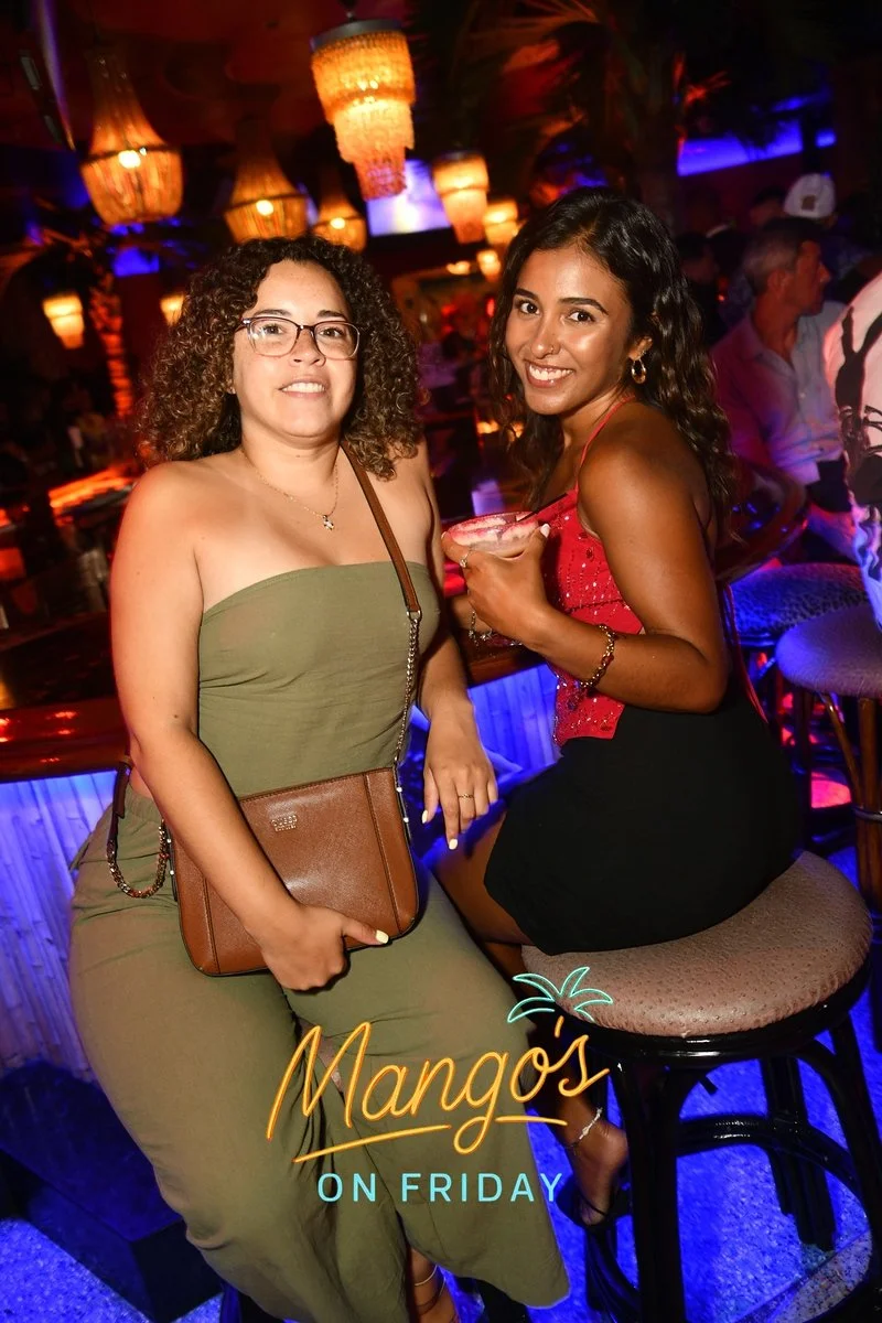 Two women at a bar or nightclub, smiling and posing for a photo. One woman is standing and wearing a strapless green dress with glasses and a brown purse. The other woman is sitting on a stool, wearing a red top and a black skirt, holding a cocktail.