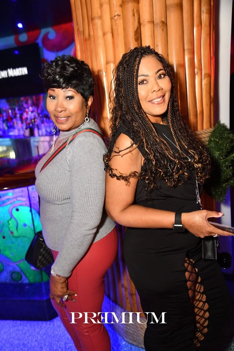 Two women standing back to back in a bar or lounge setting, smiling at the camera. One woman has short black hair and is dressed in a grey top and red pants, while the other woman has long braided hair and is wearing a black outfit with laced-up pants.