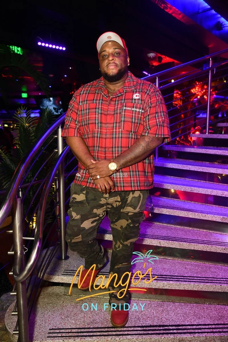 A man in a red plaid shirt and camouflage pants standing on a staircase in a neon-lit venue with 'Mangos on Friday' text at the bottom.