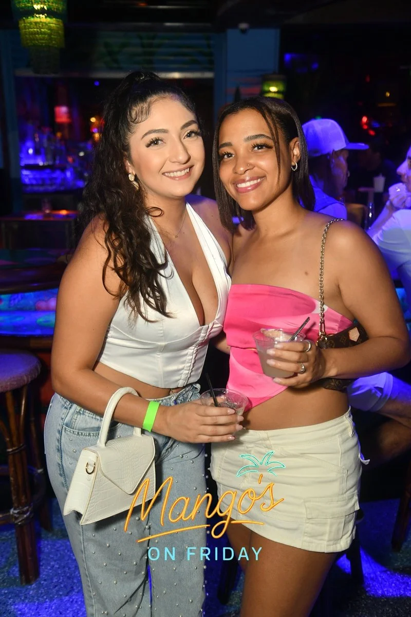 Two women smiling and holding drinks in a nightclub with colorful lighting, with a sign that says 'Mango's on Friday' at the bottom.