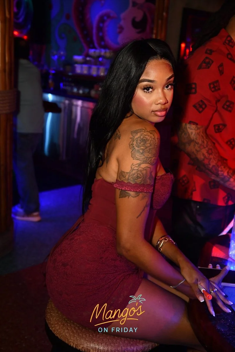 A woman with long black hair and tattoos on her arm, wearing a red off-the-shoulder dress, seated at a bar in a nightclub with colorful neon lights. The words "Mango's ON FRIDAY" are displayed at the bottom.