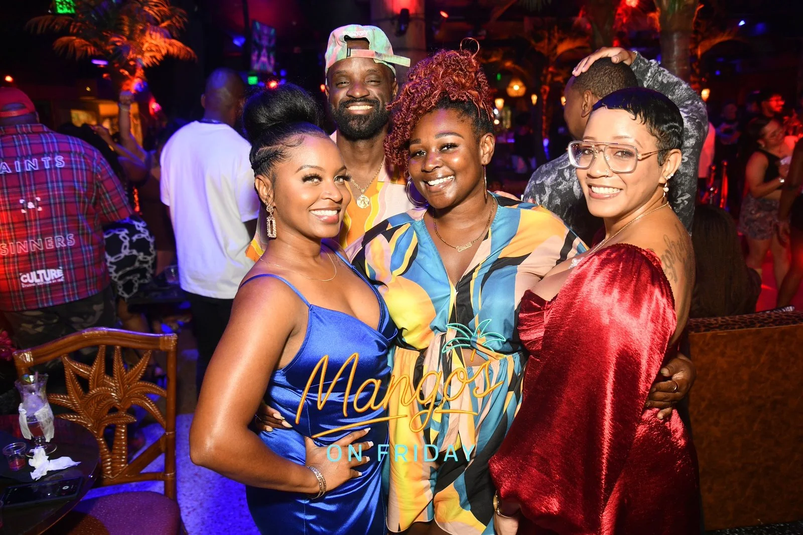 A group of five smiling people at a lively party or celebration, posing closely together. They are dressed in colorful, stylish clothing, with a vibrant, dimly lit background indicating an indoor venue with tropical or themed decor.