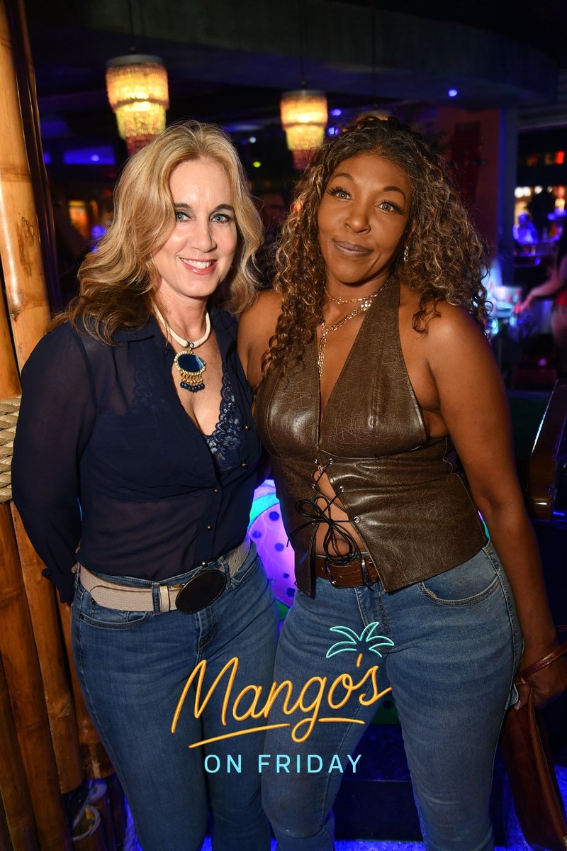 Two women standing together at nightlife bar with hanging pendant lights in background, smiling and posing for photo, with "Mango's on Friday" logo in foreground.
