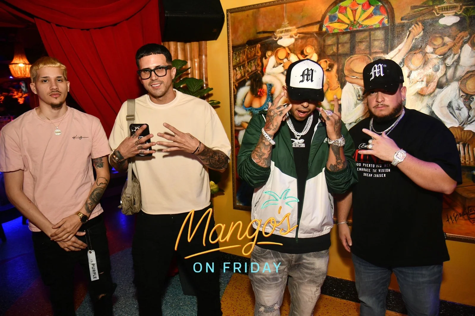 Group of four men posing at Mango's on Friday, with colorful mural behind them and neon sign in front.