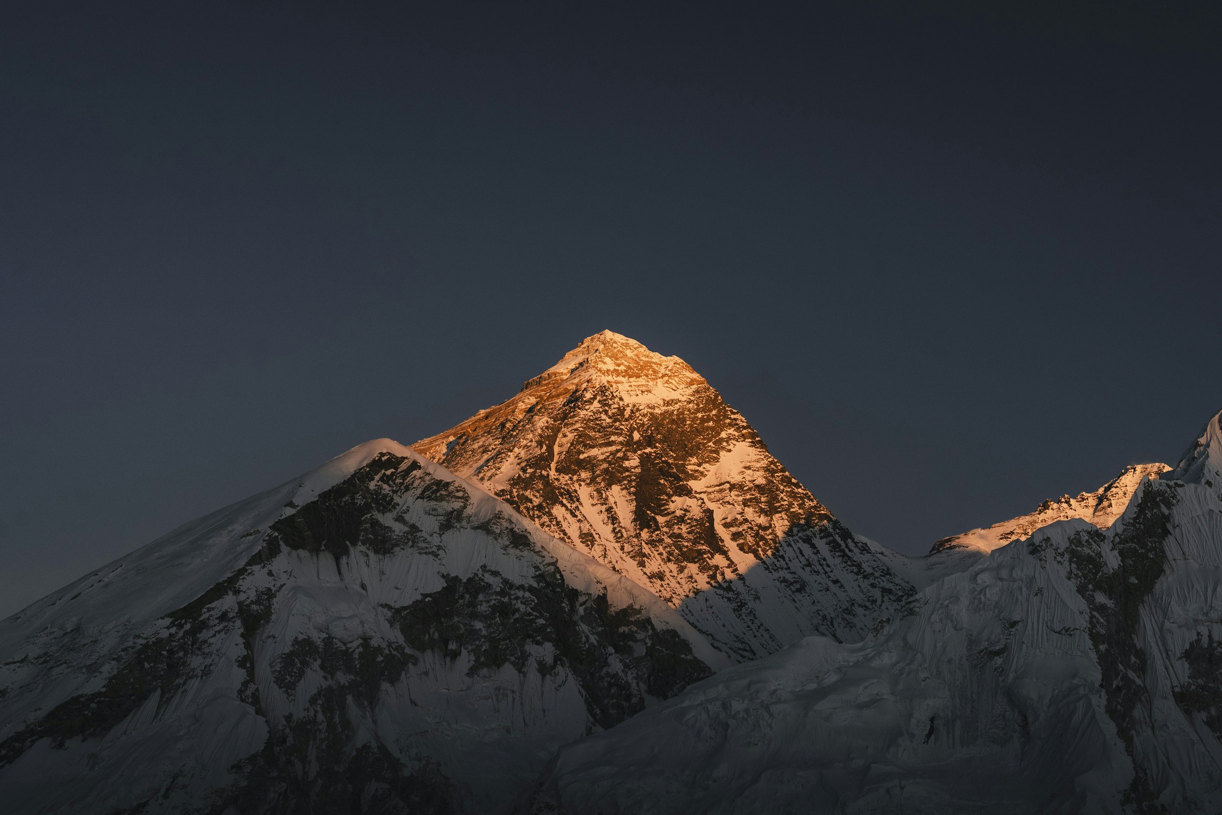 Snow-capped mountain peak illuminated by the setting or rising sun, with a dark sky in the background.