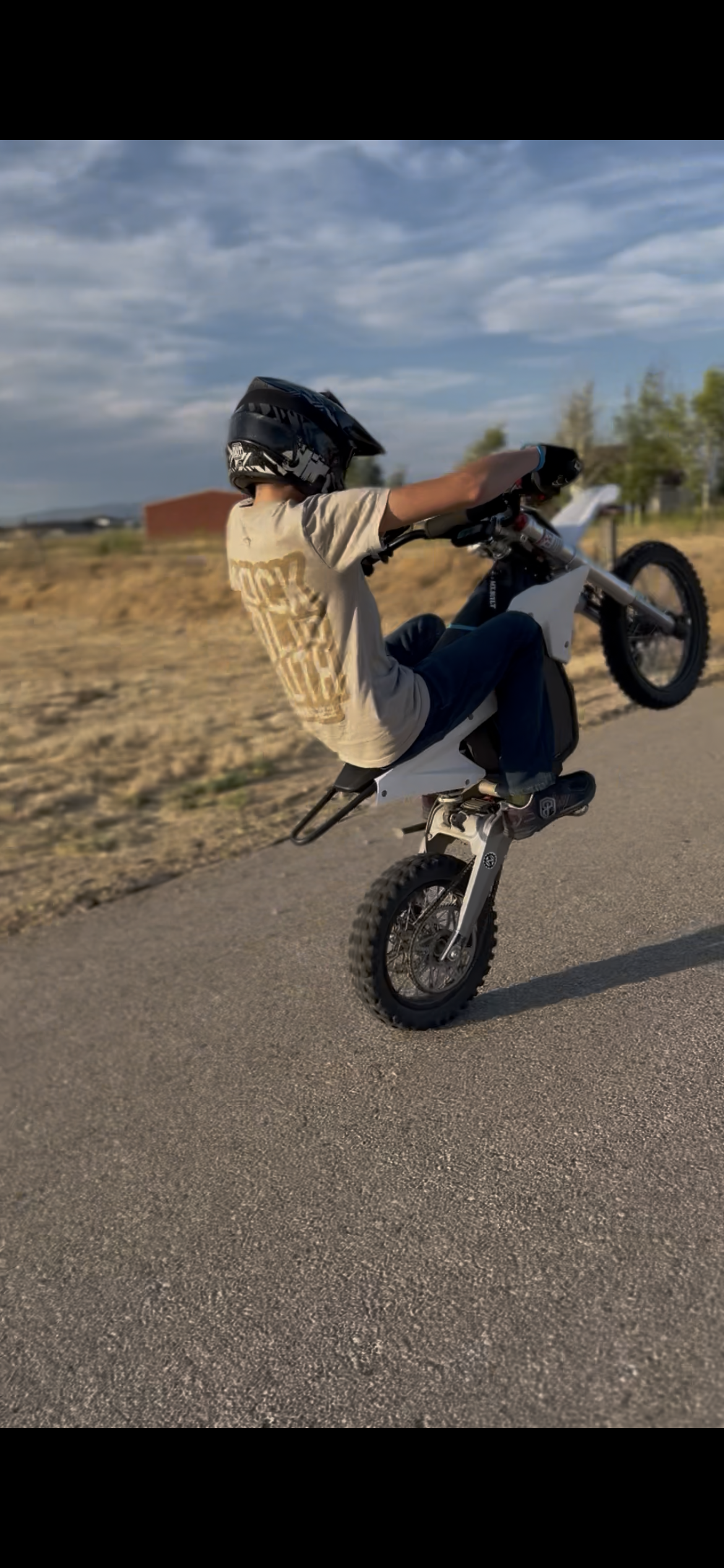 A child wearing a helmet is performing a wheelie on an electric dirt bike on a paved road in an open rural area with fields and a few trees.