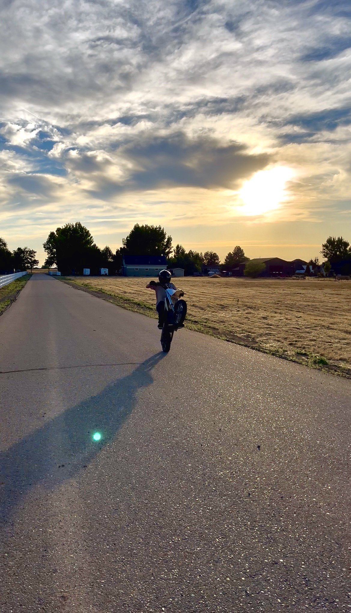 A person riding a bicycle on a paved road during sunset with a cloudy sky, trees, and houses in the background.