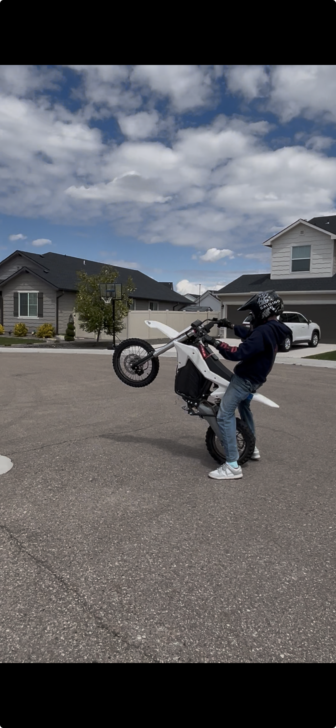 Person wearing a helmet performing a wheelie on a white electric dirt bike on a suburban street.