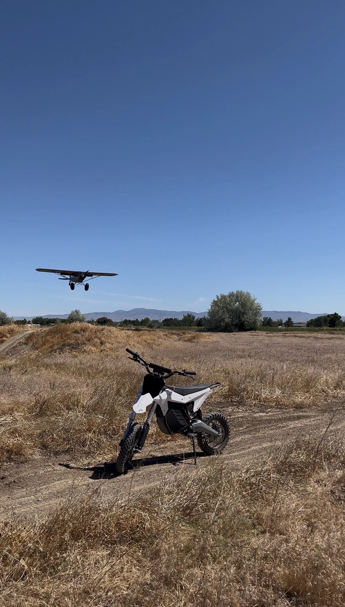 A small off-road bike is parked on a dirt path in a dry grassy field, with a mountain range in the background. A small aircraft is flying in a clear blue sky.