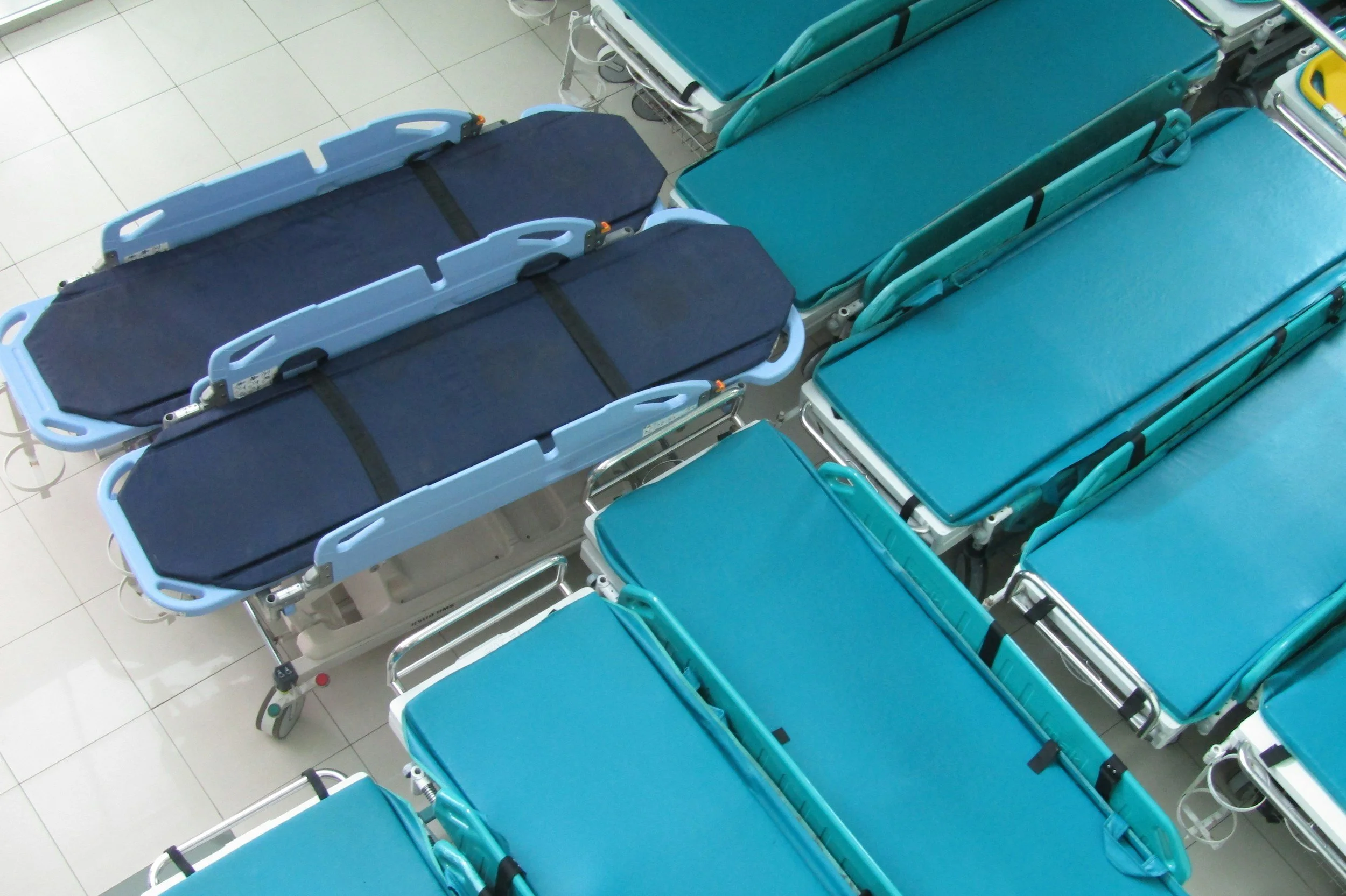 Mehrere blaue Krankenbetten in einem Krankenhausflur, einige liegen flach auf dem Boden, die Betten sind nebeneinander angeordnet.