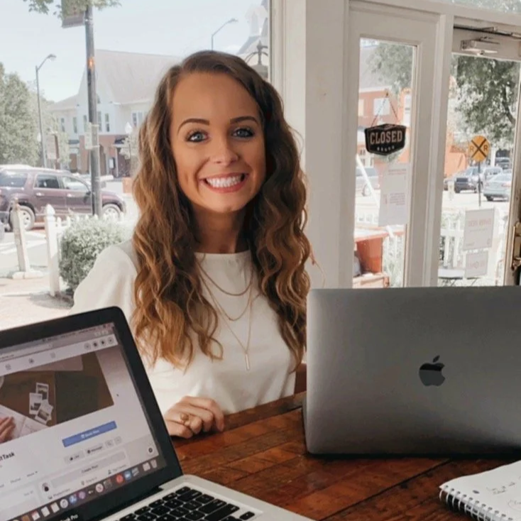 A smiling young woman with curly brown hair sitting at a wooden table in a cafe, with a MacBook and another open laptop, looking at the camera.