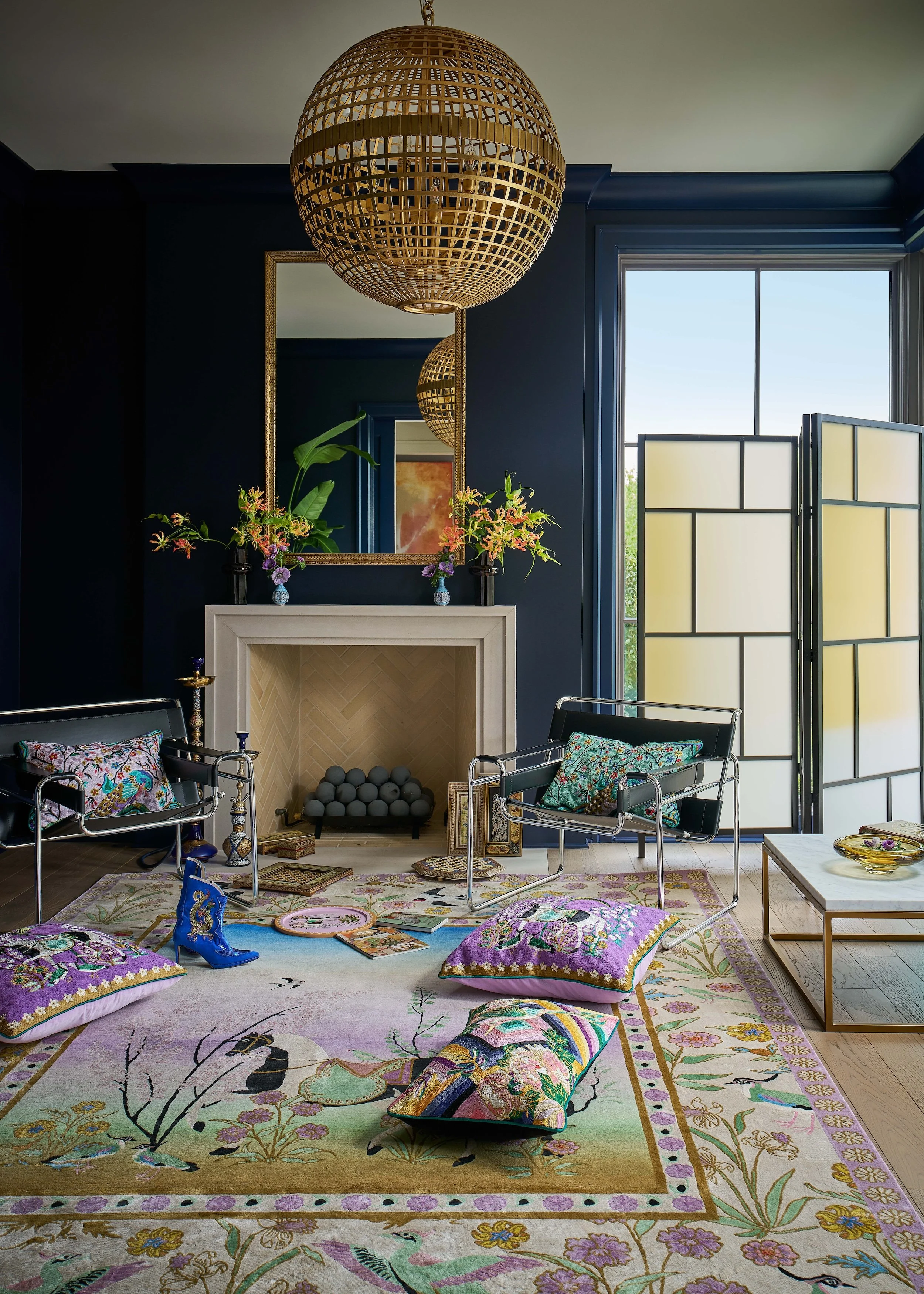 Eclectic living room with a woven globe chandelier, navy walls, ornate floral rug, fireplace, colorful pillows, modern leather chairs, and folding screen